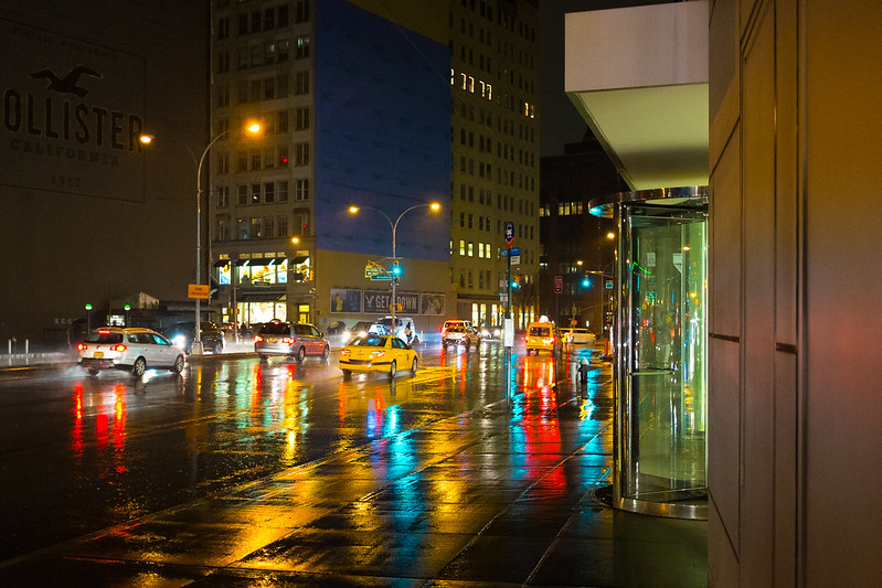 Cena noturna de Nova York com ruas molhadas, reflexos coloridos e táxis.